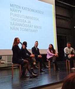 Panelistit istumassa vierekkäin lavalla, etummainen nuori puhumassa yleisölle mikrofoni kädessä. Taustalla näkyy oppilaiden keksimä kysymys: miten katsomuksesi näkyy pukeutumisessa, tavoissa ja säännöissä? Miksi näin toimitaan. 