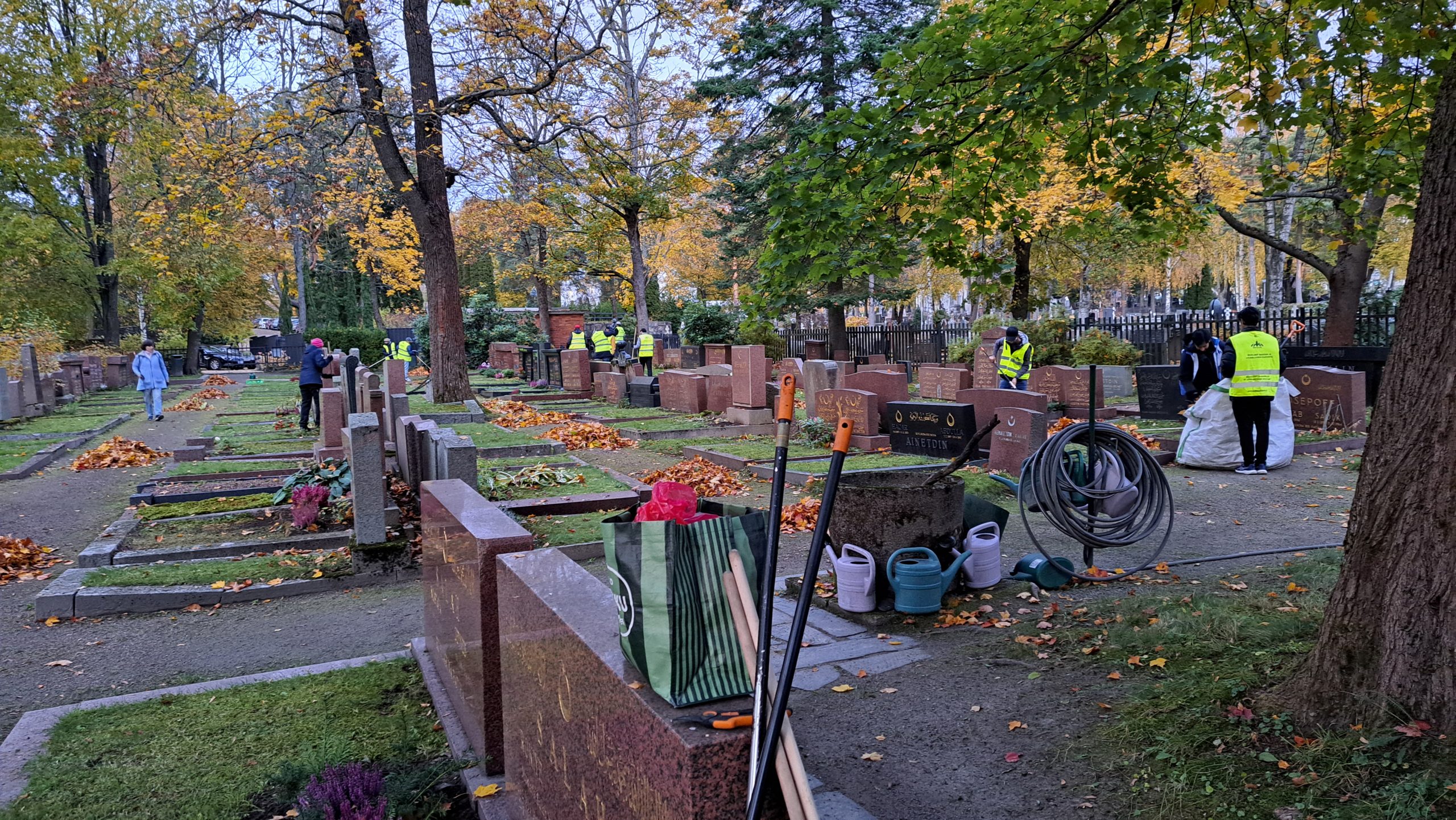 Viileässä ilmassa hautausmaata siivoavia ihmisiä. Haravoita nojaamassa hautakiveen. Puhtaaksi haravoidut haudat.
