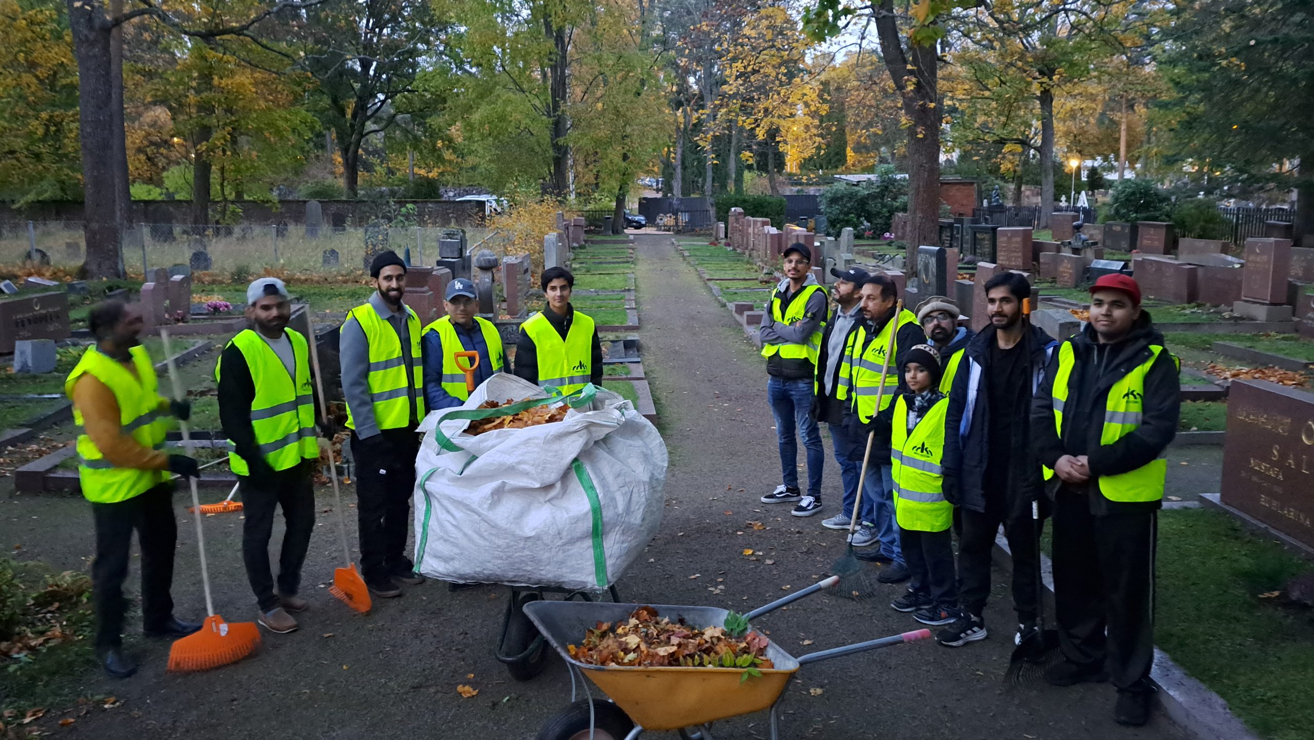 Haravoitu ja siistitty hautausmaa sekä ryhmä viherliivisiä, vapaaehtoisia siivoajia Ahmadiyya-yhteisöstä. Ihmisten keskellä kassillinen ja kottikärryllinen lehtiä.